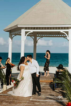 Married couple kiss on the boardwalk with crowd and gazebo in the background of a destination wedding on the beach in Mexico