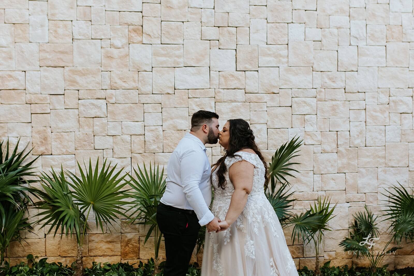 Happy couple kissing in front of a stone wall with tropical greenery.