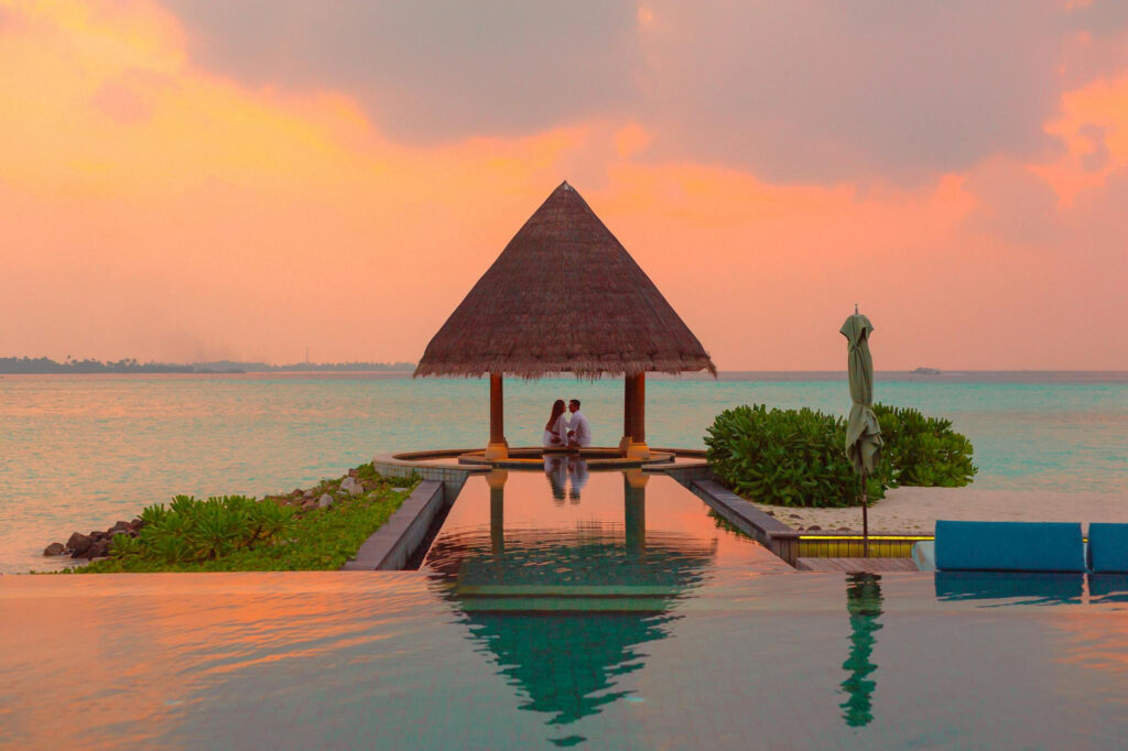 Couple sitting under a thatched pavilion by the ocean at sunset, reflected in an infinity pool.