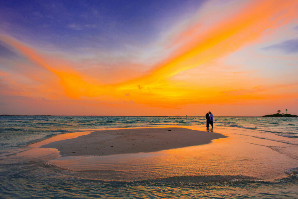 Couple embracing on a sandbar at sunset with vivid orange and purple skies over the ocean.
