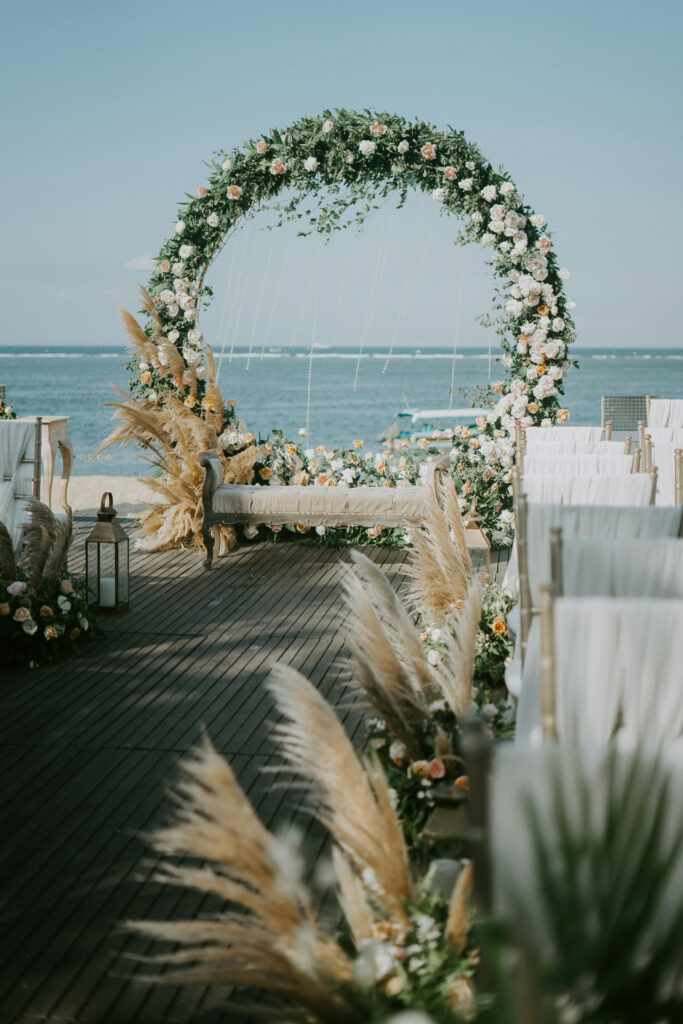 Elegant seaside wedding ceremony setup with floral arch, pampas grass, and white-covered guest chairs.