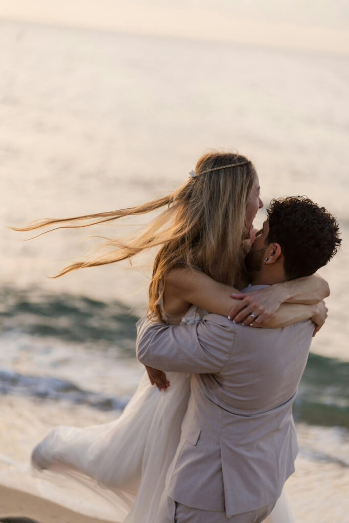 Joyful couple embracing by the ocean during a romantic beach wedding at sunset.