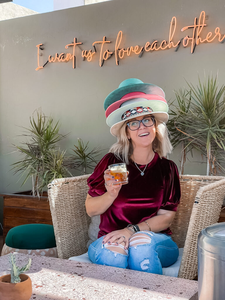 Smiling woman wearing stacked hats, sipping a drink, showcasing playful side of a Destination Wedding Planner’s travels.