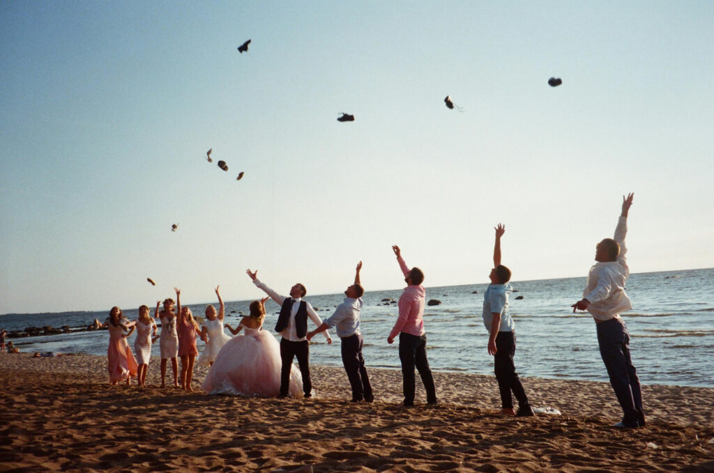 Wedding party celebrating on the beach at sunset, joyfully tossing shoes into the air.