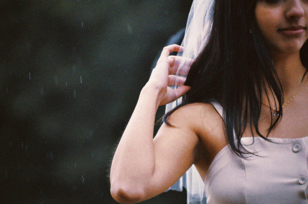 Bride brushing hair back in soft rain, wearing a veil and sleeveless dress with a gentle smile.