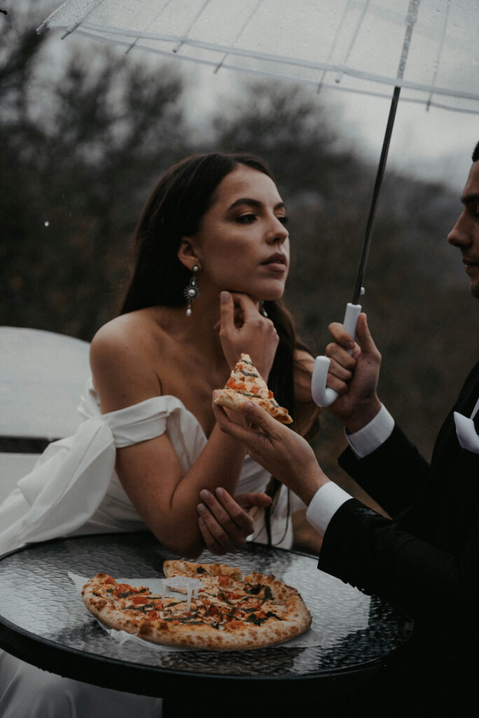 Bride enjoying pizza under umbrella with groom—playful, moody moment during a wedding in rainy season.