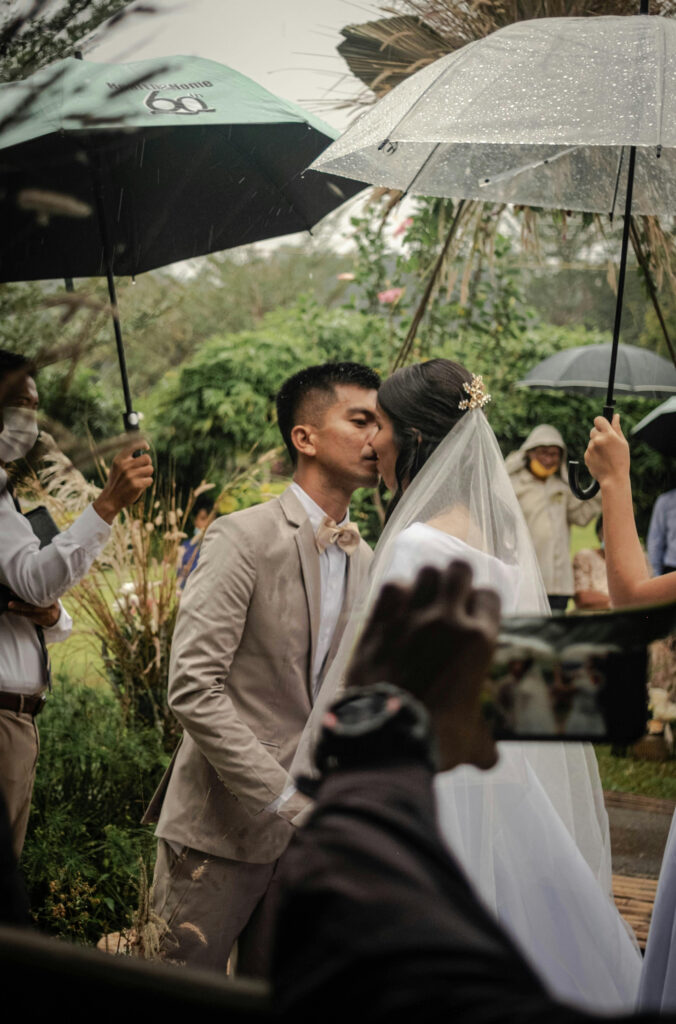 Bride and groom share a kiss under umbrellas during a romantic rainy outdoor wedding ceremony.