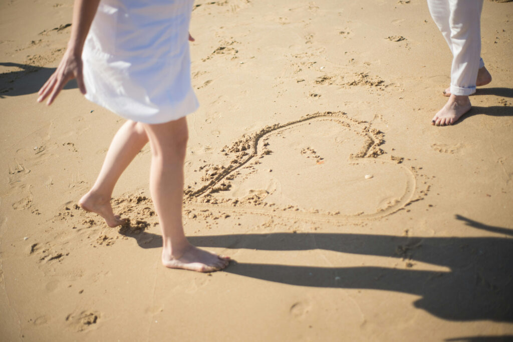 Couple walking barefoot on sandy beach near heart drawing, capturing romance and honeymoon upgrades by the sea.