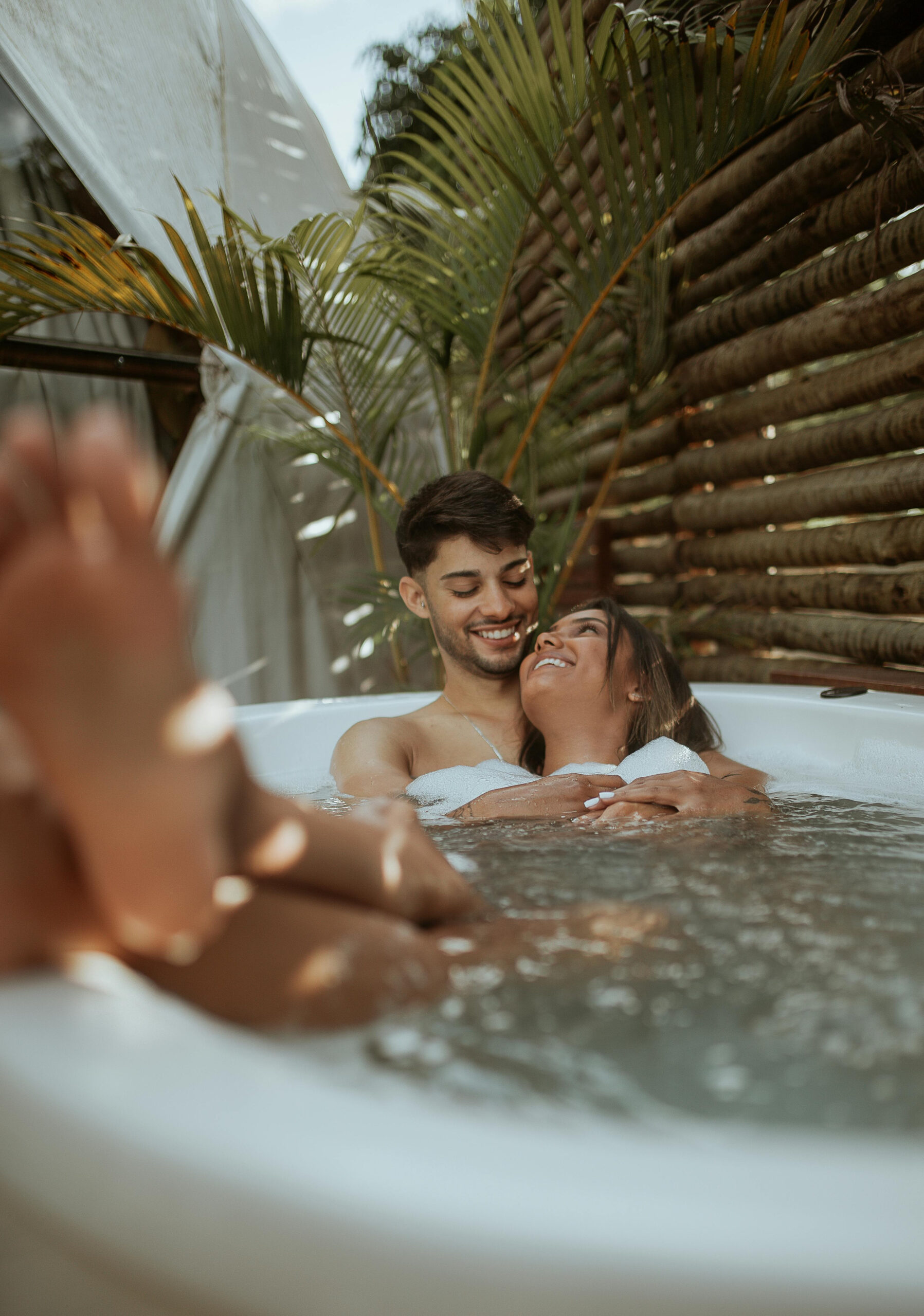 Couple enjoying a romantic wedding retreat, relaxing together in a private outdoor hot tub surrounded by tropical plants and natural wood decor.