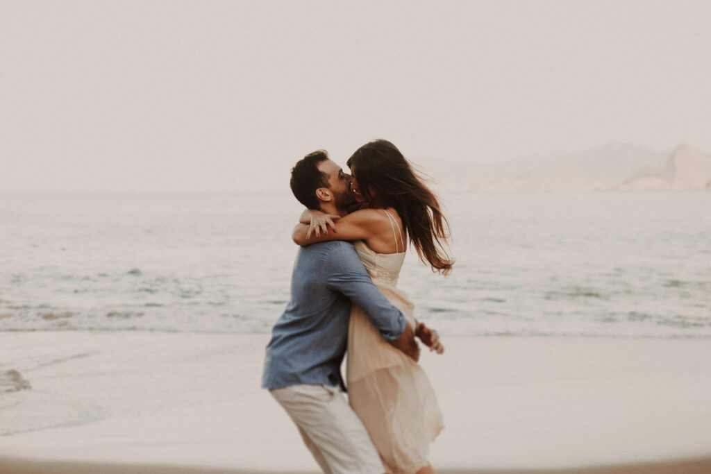 Couple embracing on a beach at sunset, with soft waves and distant mountains in the background.