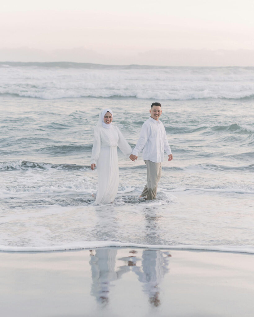 Smiling couple dressed in white walks hand in hand along the shoreline during a beach wedding moment.