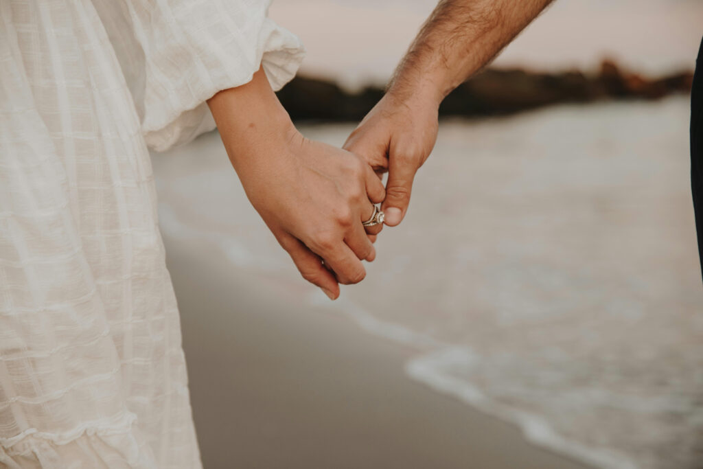 Close-up of couple holding hands on the beach, with wedding rings and shoreline softly in view and wedding ritual ideas