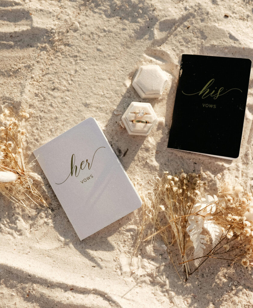 His and her vow books with wedding rings and dried florals arranged on sunlit beach sand.