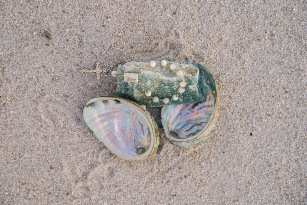 Rosary and polished green stone resting inside abalone shells on the sand, symbolizing spiritual tradition and wedding ritual ideas