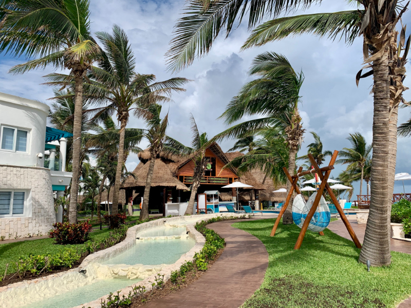 Tropical resort pathway lined with palm trees, lounge chairs, swings, and a thatched-roof beach bar.
