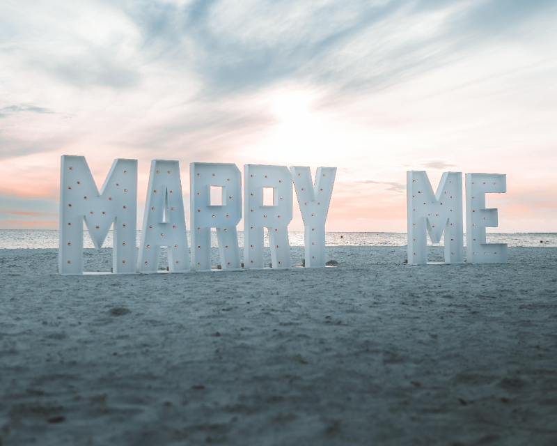 Large “Marry Me” sign on the beach at sunset, perfect for planning your 2027 wedding proposal.