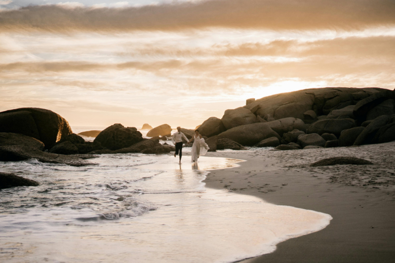 Bride and groom walking along a rocky beach at sunset, holding hands with waves at their feet.