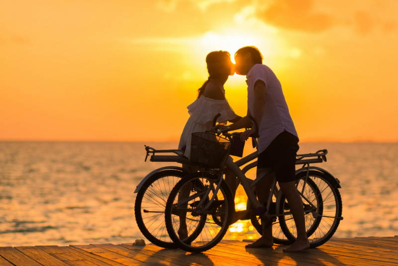 Couple kissing at sunset with bikes by the ocean, embracing love and avoiding honeymoon travel mistakes.