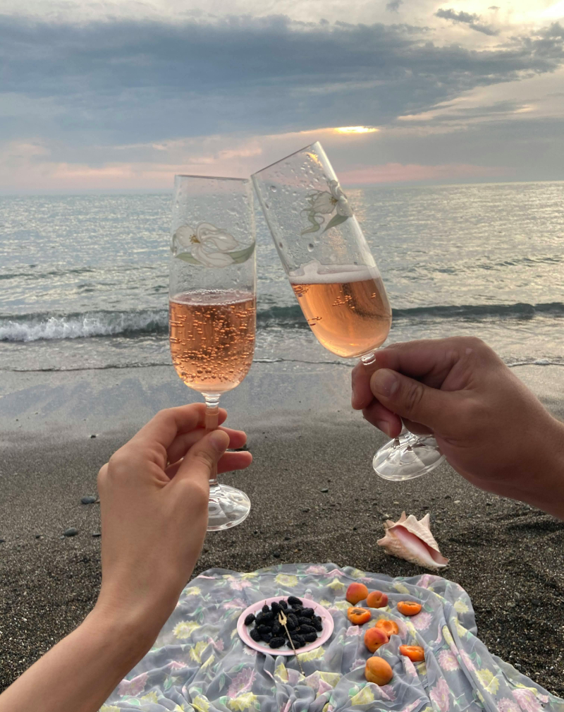 Two hands clink glasses of rosé at a beach picnic with fruit, seashells, and a sunset view.