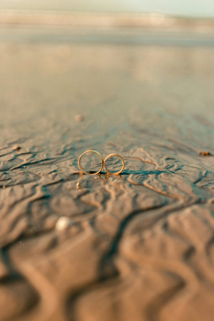 Two gold wedding rings rest on wet sand, reflecting soft light at low tide on a quiet beach.