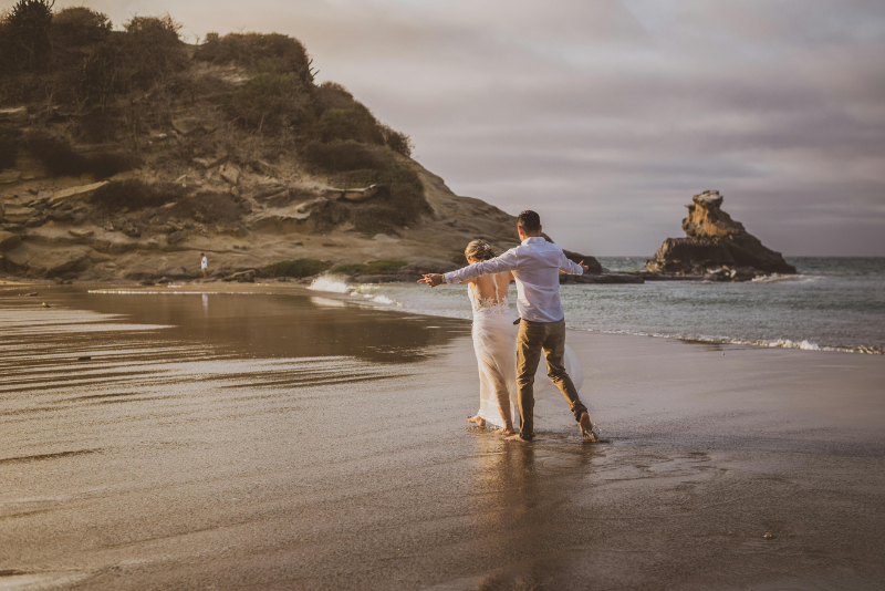 Couple dancing barefoot on a beach at their perfect destination wedding spot with rocky cliffs nearby.