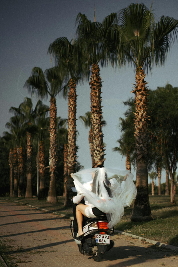 Bride on scooter with flowing veil, riding through palm trees after a luxe destination wedding.
