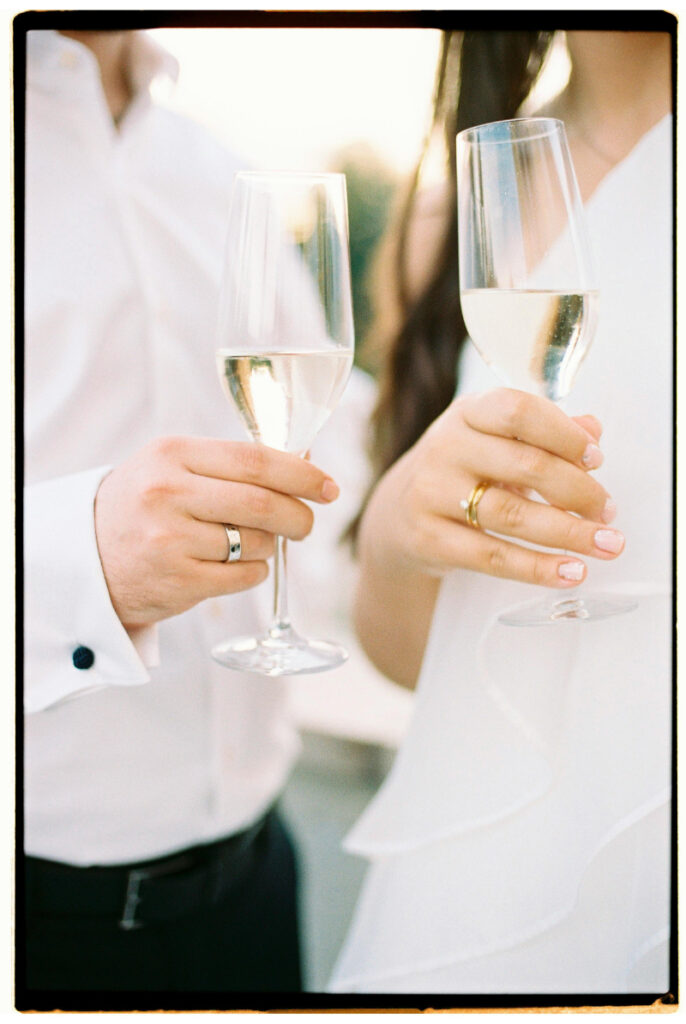 Close-up of newlyweds clinking champagne glasses, showing their wedding rings and celebration attire.