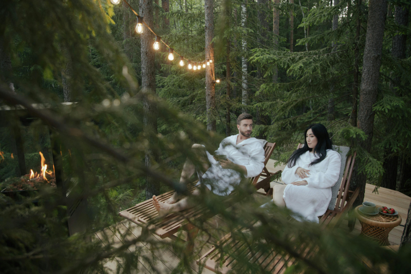 Couple relaxing in robes on a forest deck with string lights and a fire, enjoying a peaceful retreat.
