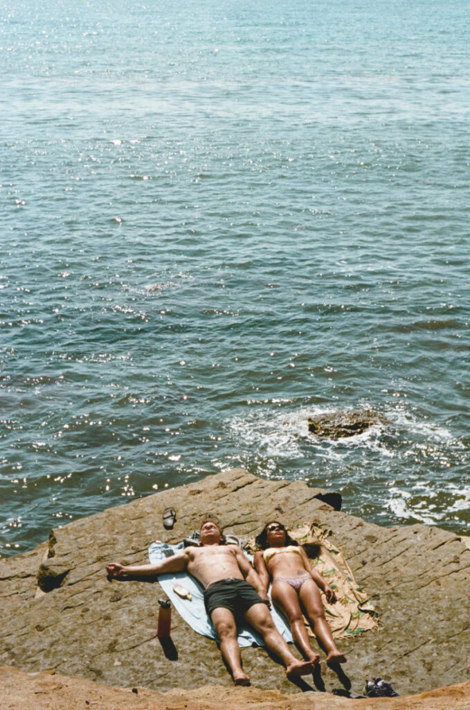 Couple sunbathing on a rocky shoreline beside the ocean, enjoying a laid-back honeymoon moment together.