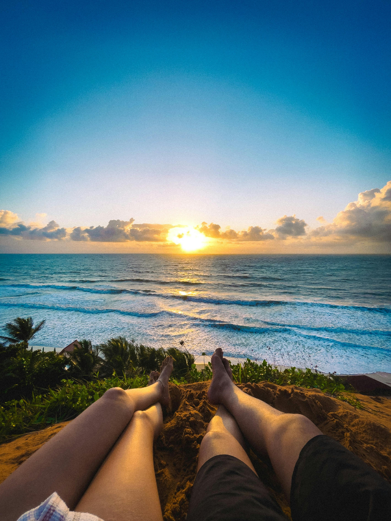 Couple relaxing on a sandy hill at sunset, overlooking ocean waves and tropical greenery below.