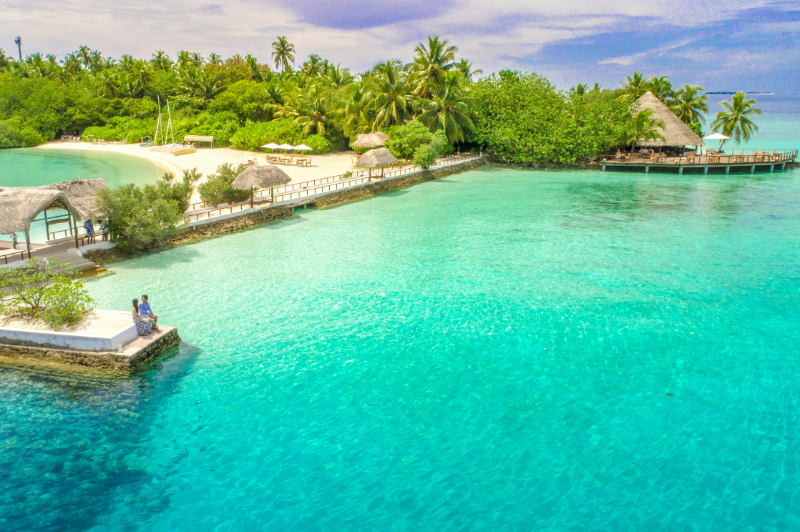 Tropical lagoon with clear turquoise water, white sand beach, and lush palm trees under a sunny sky.