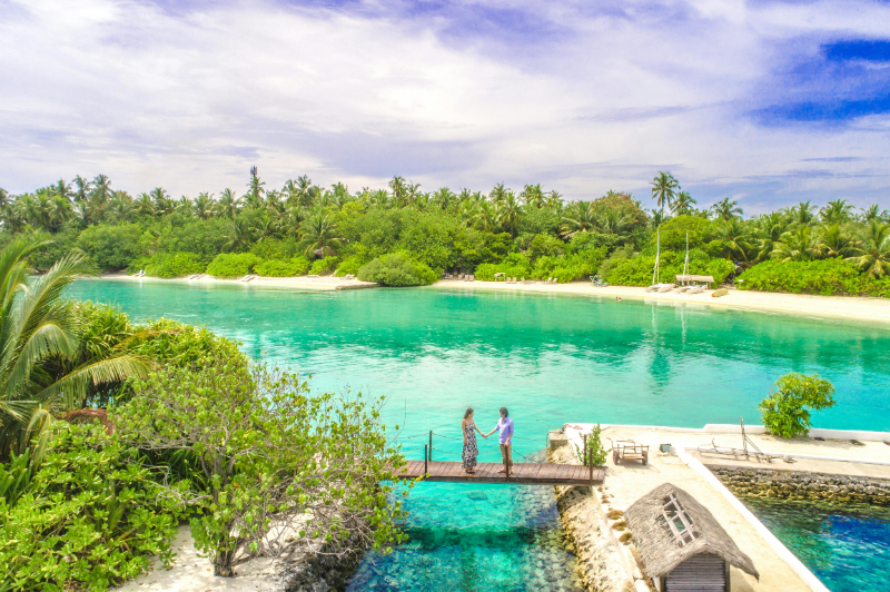 Couple holding hands on a dock over turquoise water, surrounded by lush tropical island scenery.