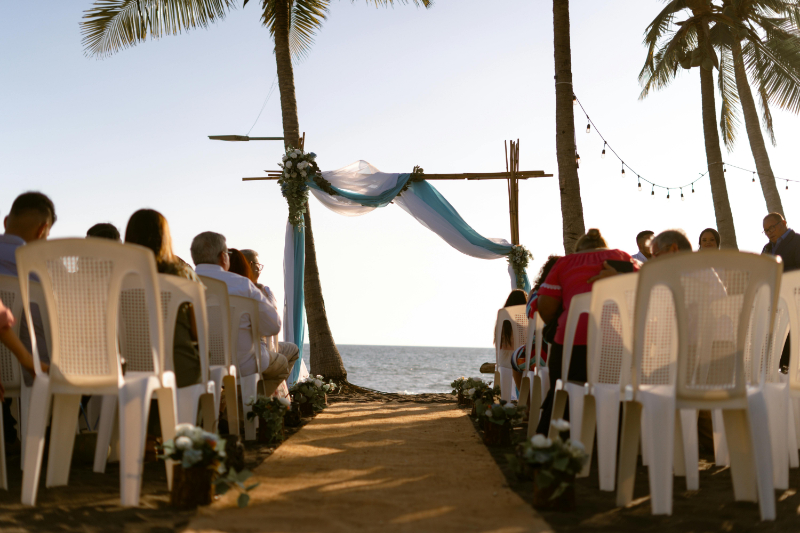 Guests attending a destination wedding seated along a beachfront aisle at sunset, facing a simple bamboo arch with flowing blue and white fabric.