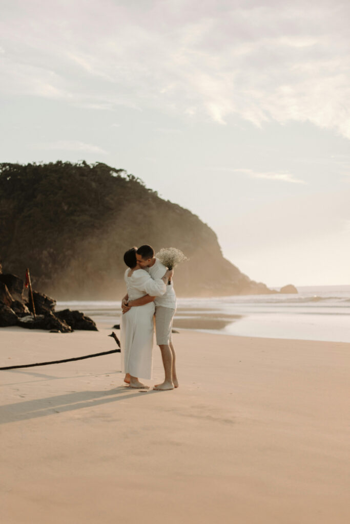 Couple embraces on a quiet beach at sunset, holding flowers with cliffs and ocean in the background.