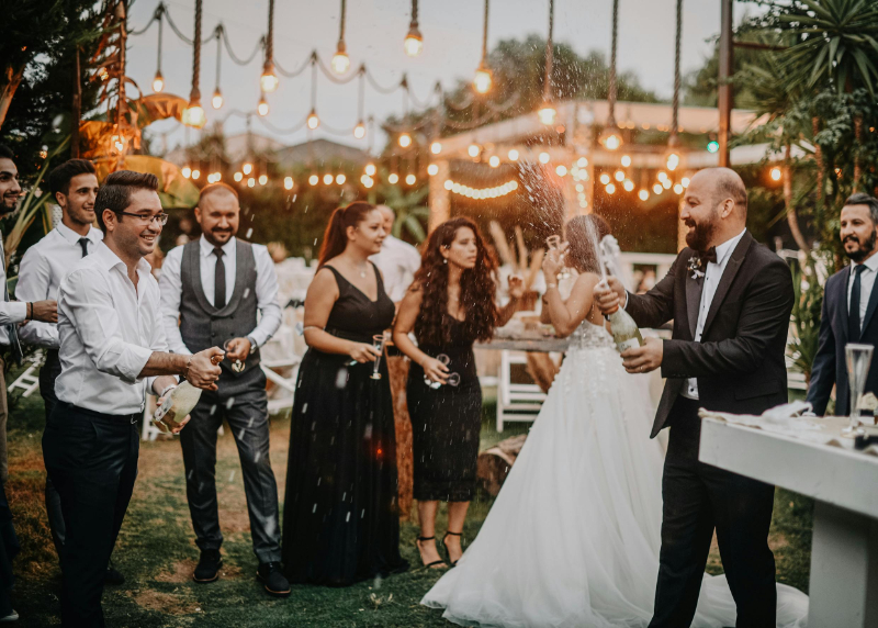 Guests attending a destination wedding celebrate outdoors with champagne, as the couple and friends gather under string lights for a lively reception.