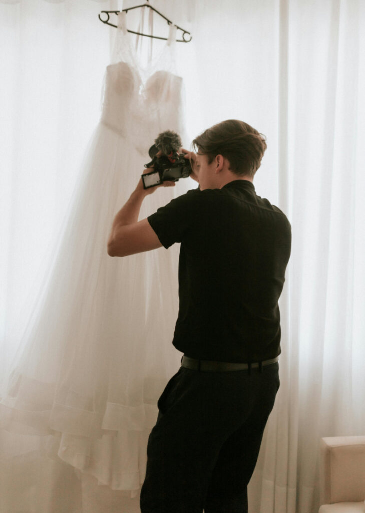 Videographer capturing bridal gown detail behind the scenes of a destination wedding near soft window light.
