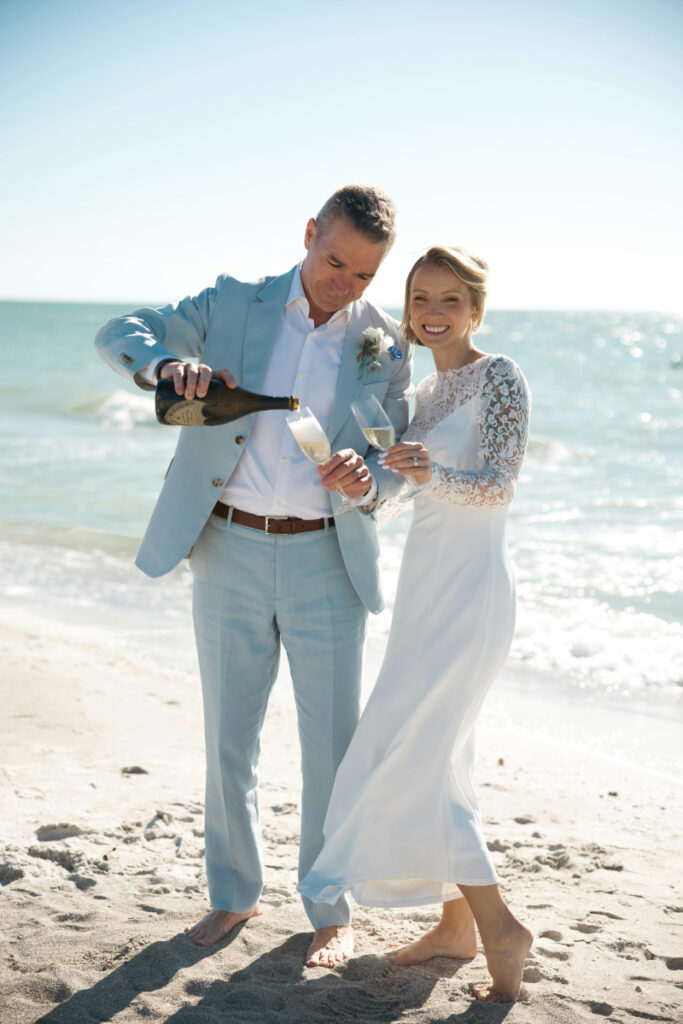 Groom pouring champagne as bride smiles beside him during a beachside wedding celebration by the ocean.