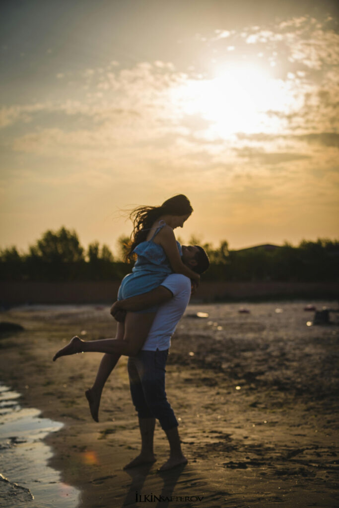 Man lifting woman at sunset with a joyful embrace on a quiet beach or shoreline - How to Pick the Right Type of Honeymoon