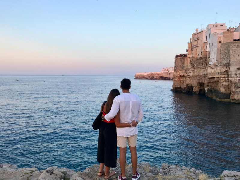 Couple embracing on rocky coastline overlooking calm blue sea at sunset.
