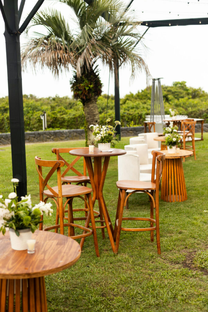Outdoor wedding cocktail hour setup with wooden high top tables, chairs, and small floral centerpieces on grass.
