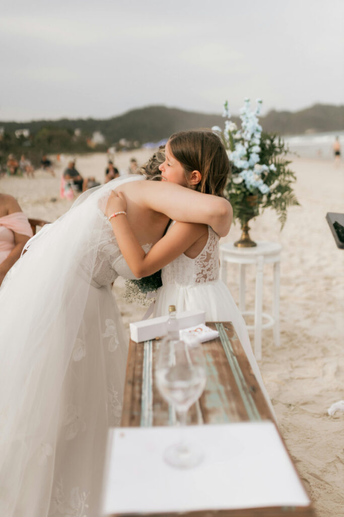 Bride hugging young girl during emotional beach ceremony moment, joyful destination wedding planning checklist inspiration.