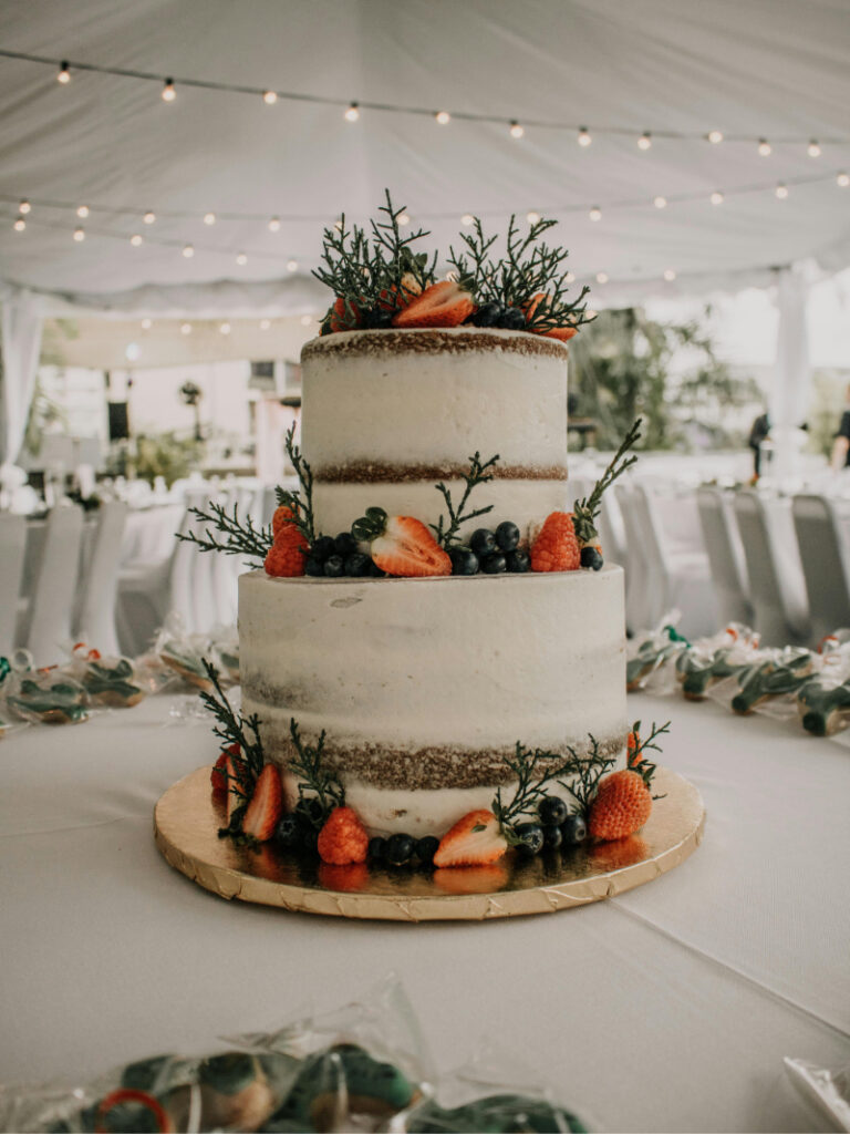 Rustic two tier wedding cake with fresh berries and greenery under reception tent string lights.