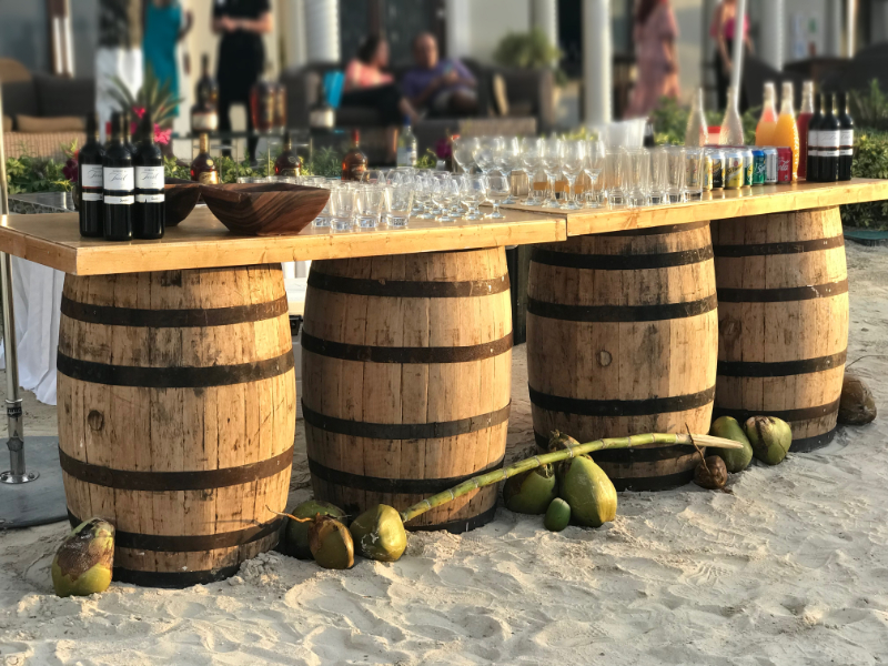 Rustic beach bar setup with wooden barrels, glassware, and fresh coconuts.