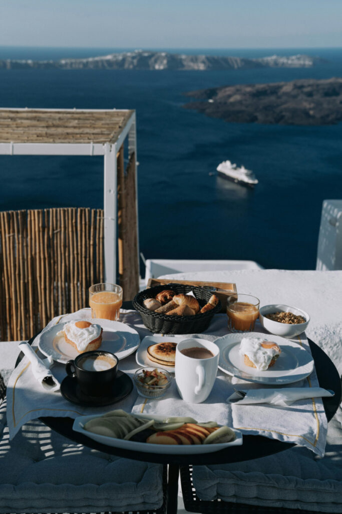 Breakfast spread with pastries, fruit, and coffee overlooking a scenic waterfront terrace.
