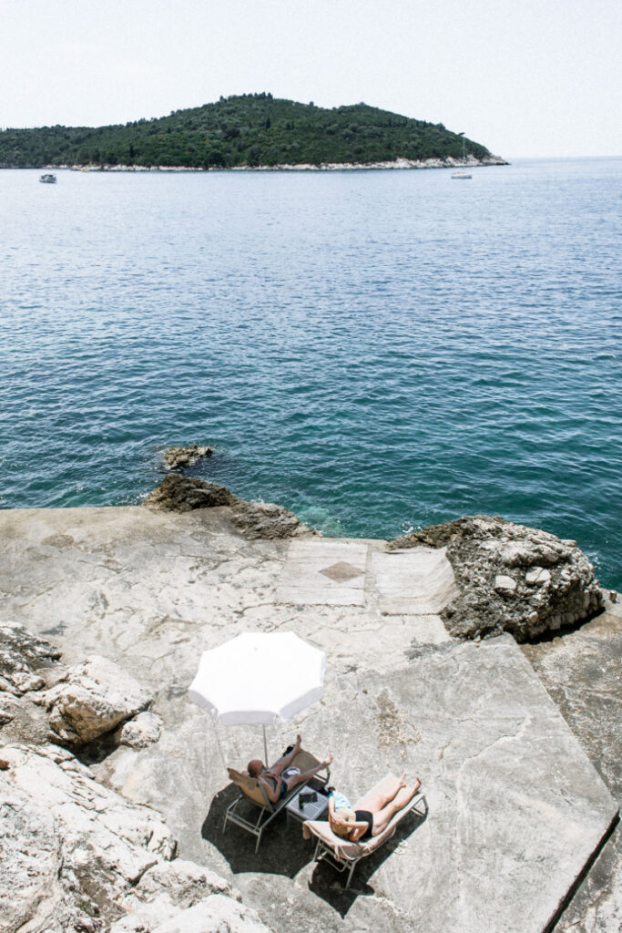Couple relaxing on lounge chairs beside a rocky coastline with clear blue water and a shaded umbrella.